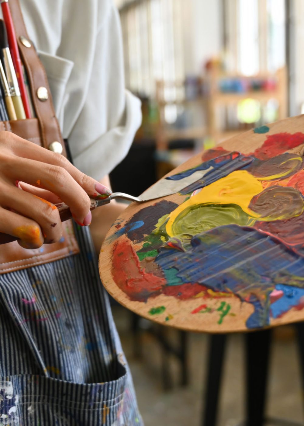 Close-up image of a young creative artist's hands smeared with watercolors holding a spatula mixing color on a palette.